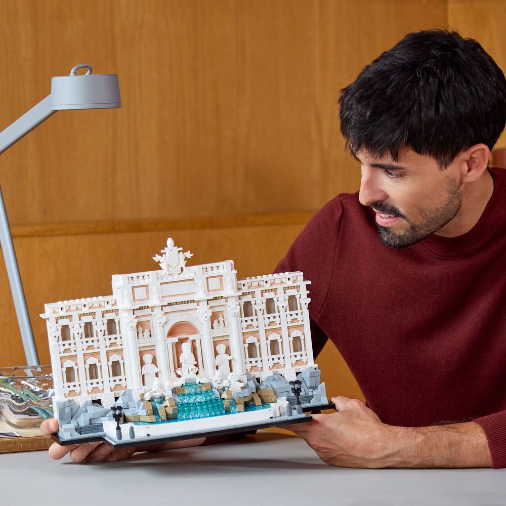 LEGO 21062 Ein Mann hält ein detailliertes LEGO-Modell des Trevi-Brunnens vor einem Holzhintergrund.